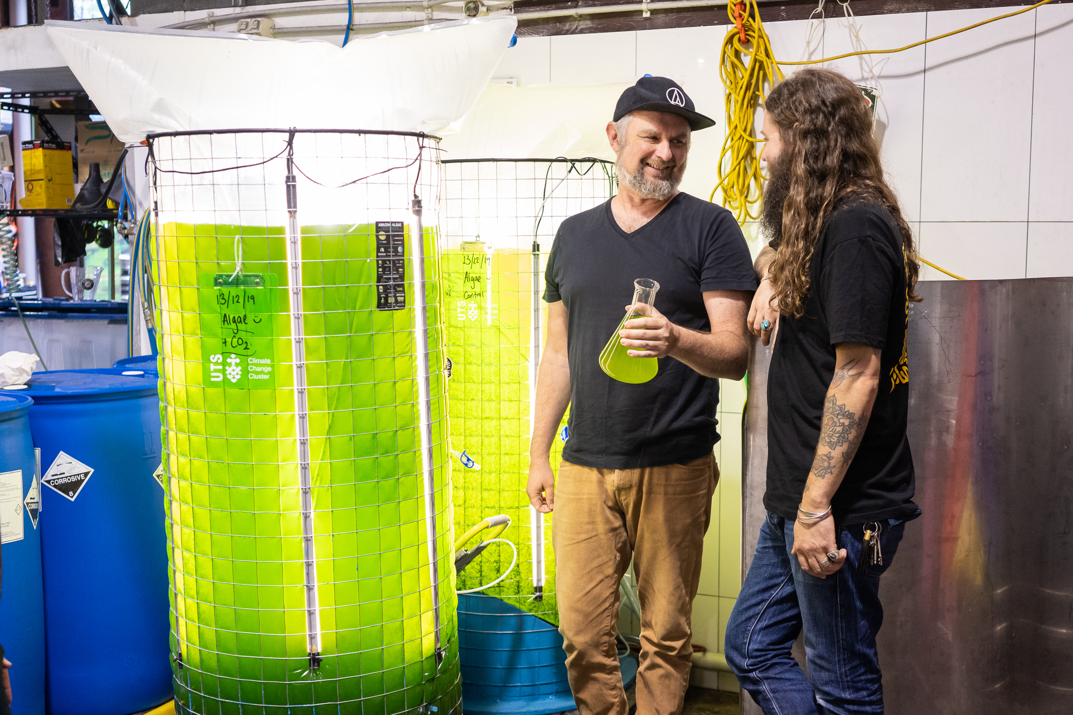 An image of the Young Henrys co-founders in their brewery, next to the 400L algae bioreactor which is glowing fluro green.