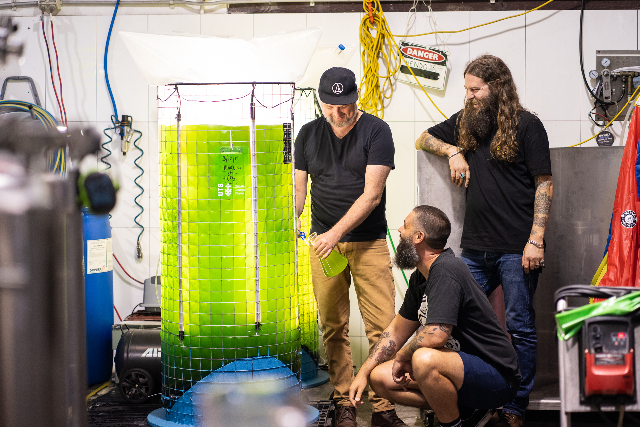 An image of the Young Henrys co-founders in their brewery, next to the 400L algae bioreactor which is glowing fluro green. 