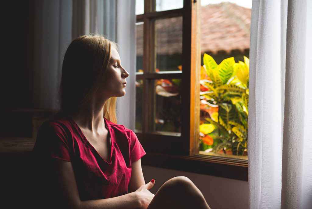 An image of a woman sitting in front of an open window with her eyes closed, meditating. 